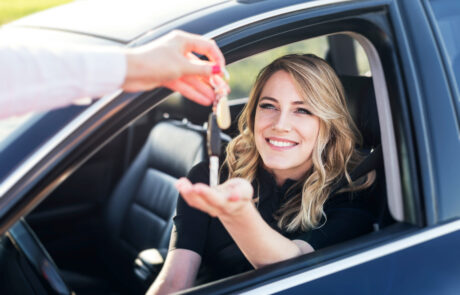 Woman sitting in driver's seat of a blue sedan holding her hand out of the open window to be handed the keys to the car.