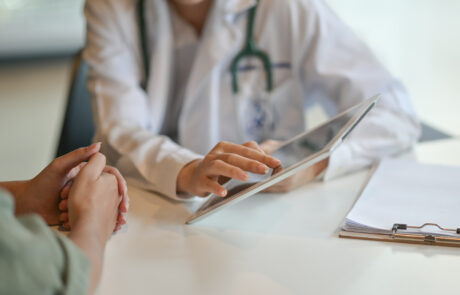 Someone speaking to a doctor. Neither person's face is visible, but you can see that one is wearing a white coat and stethoscope. That person is pointing out information on a clipboard.