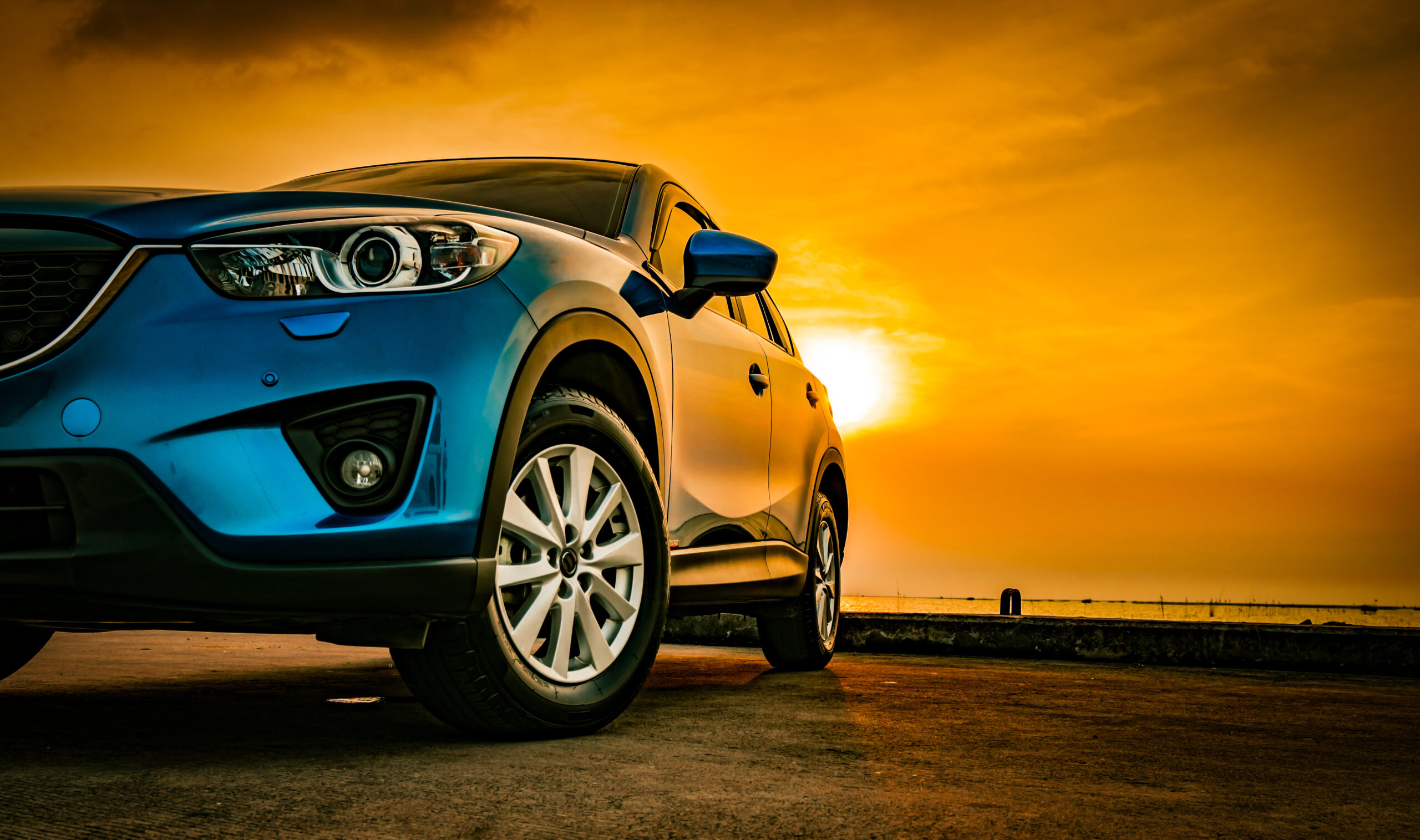 Close-up of a blue SUV parked on a paved surface at sunset, with the sun low in the sky casting a warm orange and yellow glow across the horizon.
